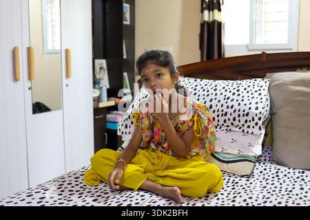 Ein Kind sitzt auf einem Bett und isst einen Snack, während er sich in einem Haus umsieht. Stockfoto