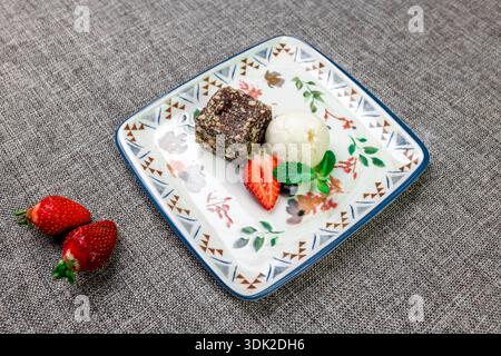 Eine Nahaufnahme eines eleganten Desserttellers mit einem Stück reichhaltigen Schokoladenkuchen (Brownie), einer Schaufel Vanilleeis und einer ganzen, in Scheiben geschnittenen frischen Stra Stockfoto