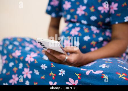 Eine Frau in einem blauen Blumenkleid sitzt auf dem Boden und schaut während der Schwangerschaft auf ihr Smartphone. Stockfoto