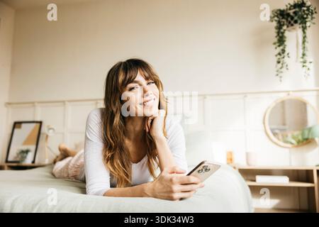 Glückliche Frau, die auf dem Bett liegt, Freizeit genießt und ihr Handy durchsucht Stockfoto