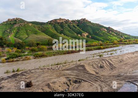 Der schlammige Fluss fließt an üppigen grünen Hügeln vorbei, die mit Felsen und verstreuten Bäumen bedeckt sind, unter einem bewölkten Himmel in der Gegend um Frank A. Miller Mount Rubidoux. Stockfoto