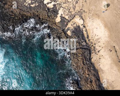 Aus der Vogelperspektive auf die wilde, zerklüftete Küste, wo das türkisfarbene Meer gegen die dunklen vulkanischen Felsen prallt, bevor es auf den weichen Sandstrand El C trifft Stockfoto