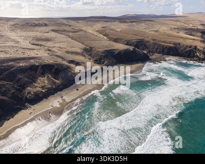 Aus der Vogelperspektive der türkisfarbenen Wellen, die gegen die Sandküste prallen und auf die trockene Landschaft treffen, Playa de la Pared, Fuerteventura, Spanien. Stockfoto