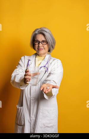 Eine leitende Ärztin, die eine Pille und ein Glas Wasser auf gelbem Hintergrund anbietet Stockfoto