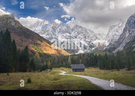 Blick aus der Vogelperspektive auf einen gewundenen Pfad, der zu einer rustikalen Hütte führt, eingebettet in das vom Herbst geküsste Laub und die schneebedeckten Gipfel, Kranjska Gora, Jesenice, Slove Stockfoto