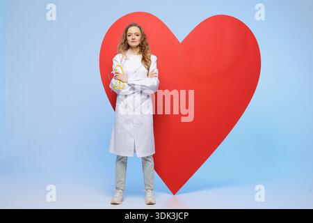 Eine Ärztin im weißen Mantel steht selbstbewusst vor dem großen roten Herzsymbol im Studio. Konzeptbild für kardiologische Sensibilisierung Healthcare Adverti Stockfoto