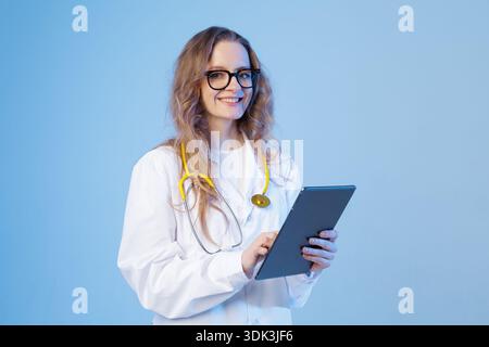 Weibliche Ärztin im weißen Mantel mit Stethoskop verwendet eine Tablette auf blauem Studiohintergrund. Sauberes professionelles Image für Gesundheitstechnik, medizinische Nachteile Stockfoto
