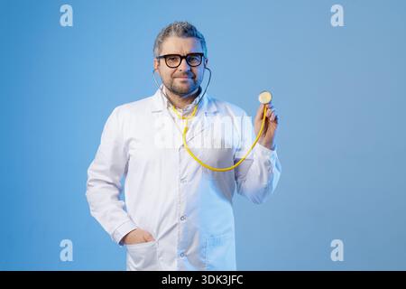 Männlicher Arzt im weißen Mantel hält ein Stethoskop vor blauem Atelierhintergrund. Sauberes professionelles Image für Gesundheitsbewusstsein, medizinische Untersuchung Stockfoto