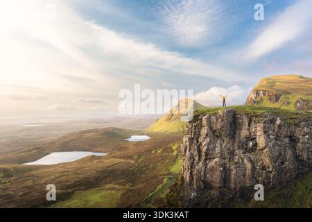 Aus der Vogelperspektive erhebt sich triumphierend auf einer zerklüfteten Klippe, die Arme zum Himmel erhoben, mit Blick auf die weite Landschaft mit sanften Hügeln und Seren Stockfoto