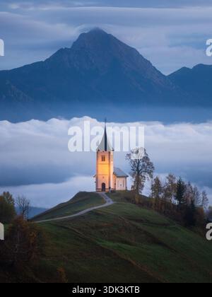 Blick aus der Vogelperspektive auf die beleuchtete Kirche St. Primus und Felicianus inmitten eines Meeres von Morgennebel vor der Kulisse majestätischer Berge, Jamnik, Slove Stockfoto