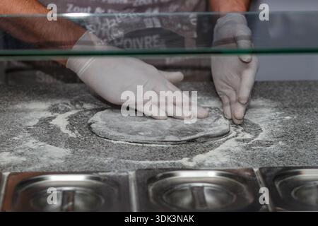 Koch Hände in weißen Handschuhen kneten frischen Pizzateig auf bemehltem Granittisch in der kommerziellen Küche. Stockfoto