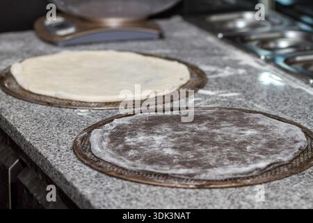 Zwei rohe Pizzateigplatten auf Metallplatten auf Granittisch, bereit für Tomatensoße. Stockfoto