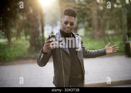 Afrikanischer Mann, der spielerisch ein Selfie auf einer Straße vor der Natur des Parks macht, Sonnenbrille und Lederjacke trägt, Stil, stilvoll, cool Stockfoto