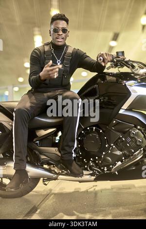 Modischer Mann sitzt auf Motorrad, afrikanischer Mann mit Sonnenbrille sitzt zuversichtlich auf einem Motorrad drinnen, Trend, Shopping Motorrad, Biker, Moto Stockfoto