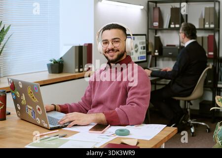 Junger Erwachsener Mann aus dem Nahen Osten lächelt, während er im modernen Büro einen Laptop benutzt Stockfoto
