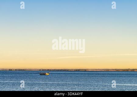 Während das erste Licht der Dämmerung durch den feuchten Dunst bricht, ruht ein einsames Boot auf einer riesigen, tiefblauen Weite. Das ruhige Wasser spiegelt den goldenen Himmel wider, c Stockfoto