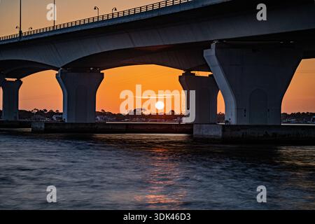 Die untergehende Sonne brennt ein Loch in den orangefarbenen Himmel, perfekt eingerahmt unter den steifen Betonbögen der Hathaway Bridge. Wenn die Dämmerung über uns herabsteigt Stockfoto