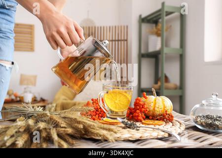 Weibliche Hand, die Tee aus der Teekanne in eine Tasse in der Nähe eines Weizenbündchens und eines ebereschen auf dem Tisch im Zimmer gießt. Nahaufnahme Stockfoto