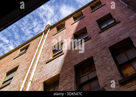 Eine alte Ziegelbauwand mit Fenstern und Abflussrohren wird aus einem niedrigen Winkel erfasst und offenbart einen wolkigen blauen Himmel darüber. Stockfoto