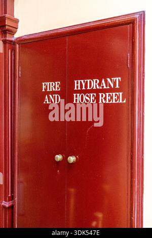 Ein roter doppeltüriger Schrank mit der Aufschrift „FIRE AND HYDRANT SCHLAUCHTROMMEL“ ist an einer hellen Wand deutlich sichtbar zu sehen. Stockfoto
