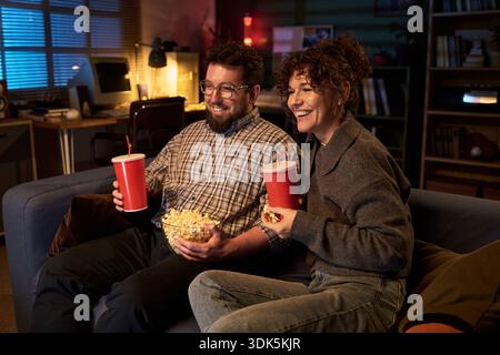 Kaukasischer Mann und kaukasische Frau sitzen auf dem Sofa und halten Popcorn und alkoholfreie Getränke lächelnd, während sie zusammen Filme im gemütlichen Wohnzimmer in der Nacht sehen Stockfoto