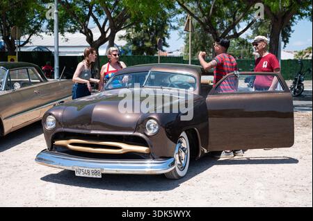 Ein Custom 1949 Ford Mercury Shoebox Hot Rod in Braun mit abgesenktem Dach und offener Tür, mit Freunden im Hintergrund, die sich in einem Oldtimer unterhalten Stockfoto