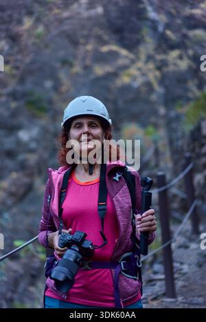 Lächelnde Wanderer mit Kamera und Helm auf dem Masca-Schlucht-Pfad zwischen vulkanischen Felswänden und Geländern Stockfoto