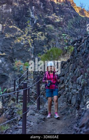 Lächelnde Wanderer mit Kamera und Helm auf dem Masca-Schlucht-Pfad zwischen vulkanischen Felswänden und Geländern Stockfoto