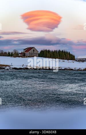 Blick auf den rauschenden Fluss, der den pastellfarbenen Sonnenuntergangshimmel und die Lentikularwolke über einer schneebedeckten Landschaft reflektiert, Tromso, Norwegen. Stockfoto
