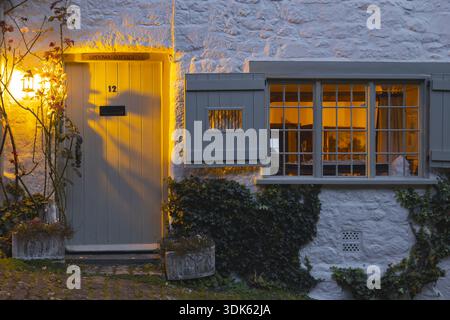 Blick auf eine malerische Cottage-Fassade leuchtet mit warmem Licht, das vom Fenster und der Tür auf die mit Efeu bedeckten Steinmauern in Shaftesbury, United Kingd ausstrahlt Stockfoto