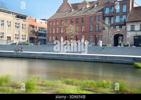 Eine helle urbane Flussszene mit Backsteinhäusern, breiten Stufen zum Wasser und Menschen, die entlang einer Uferpromenade spazieren, Radfahren und sich entspannen Stockfoto