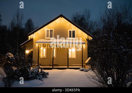 Gemütliches Haus bedeckt mit Schnee am ruhigen Winterabend mit warmen glühenden Weihnachtslichtern, umgeben von Waldbäumen und friedlicher Schneelandschaft. Stockfoto