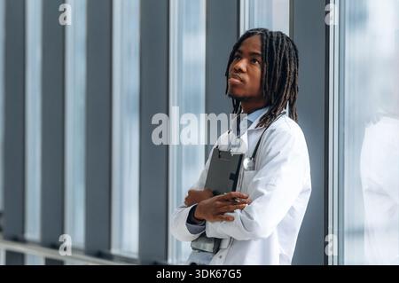 Lehnen Sie sich auf das Geländer in der Nähe von Fenstern. Der junge schwarze Doktor ist auf dem Flur. Stockfoto