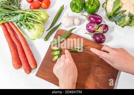 Blick von oben auf einen Koch, der Gemüse auf hölzernem Schneidebrett schneidet. Blumenkohl, Tomaten, Karotten, Auberginen, Kohlrabi, grüner Pfeffer und okr Stockfoto