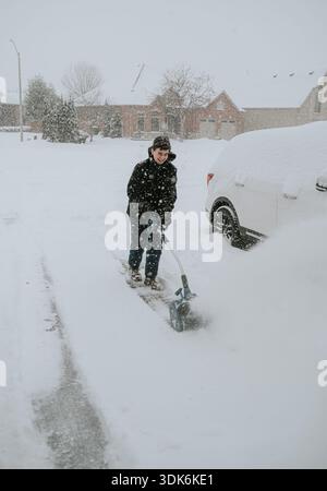 Teenager, der im Winter Schnee von der Einfahrt mit einer elektrischen Schaufel bläst. Stockfoto