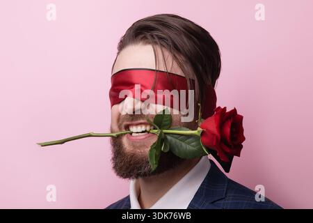Bärtiger Mann in einem Anzug mit roter Augenbinde, beißt einen roten Rosenstiel vor einem pastellrosa Hintergrund. Konzept von Romantik, Überraschung und verspielter werteliste Stockfoto