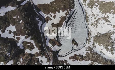 Aus der Vogelperspektive auf einen gefrorenen See, der die hellen Farben des Himmels widerspiegelt, eingebettet in die schroffen, schneebedeckten Gipfel und gewundenen Straßen der italienischen Alpen, Colle Stockfoto