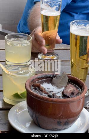 Mann mit einem Glas Bier auf dem Tisch mit Feijoada. Das bekannteste Gericht der brasilianischen Küche. Beilagen und Caipirinha, das brasilianische Getränk Stockfoto