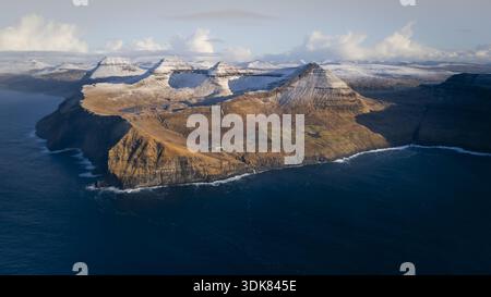 Blick aus der Vogelperspektive auf zerklüftete Klippen, die auf das tiefblaue Meer treffen, schattige Hänge im Kontrast zu schneebedeckten Gipfeln, eine ruhige Landschaft, Torshavn, Färöer Island Stockfoto