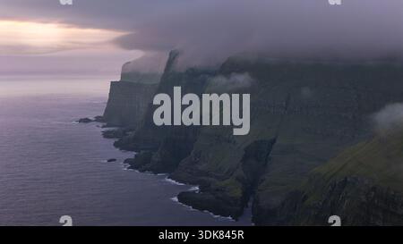 Aus der Vogelperspektive sehen Sie dramatische, in Nebel gehüllte Klippen und treffen auf das dunkle Meer unter einem pastellfarbenen Himmel, Torshavn, Färöer Inseln. Stockfoto
