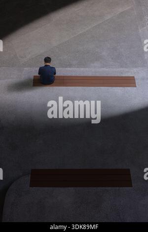 Der Blick auf eine Einzelfigur sitzt auf einer Holzbank, die in das Zusammenspiel von Licht und Schatten getaucht ist und dramatische Muster auf dem Betonboden Punggo wirft Stockfoto