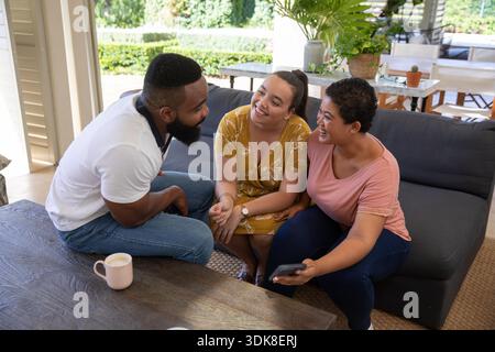 Verschiedene Freunde sitzen auf dem Sofa in der Lounge mit der Terrasse und halten ein Smartphone, während sie an einem hellrosa Becher schlürfen. Gemütlich, Freundschaft, Freizeit, Outdoor, lebhaft, lässig, Entspannung || Model veröffentlicht Stockfoto