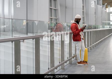Aufmerksamer afroamerikanischer Reisender mit Smartphone am Flughafenterminal, mit Gepäcktasche unterwegs. Passagier wartet auf ihren Flug, überprüft die Flugzeit online, steht im Bahnhof || Model veröffentlicht Stockfoto