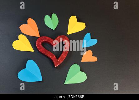 Rotes Holzherz umgeben von bunten Papierherzen auf schwarzem Hintergrund Stockfoto