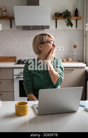 Verschlafene Geschäftsfrau mittleren Alters, gähnt und dehnt sich, sitzt am Tisch und arbeitet von zu Hause aus am Laptop. Stockfoto
