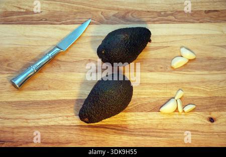 Flache Lagen frischer, reifer Avocados aus ganz Hass, die mit scharfem Küchenmesser und frischem Bio-Knoblauch auf einem hölzernen Schneidebrett angeordnet sind. Minimalistisch. Stockfoto
