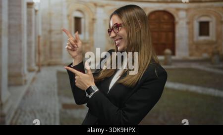 Frau zeigt beide Zeigefinger, während sie auf eine alte Straße mit Kopfsteinpflaster und Steinfassaden lächelt; fröhliches Vertrauen. Stockfoto