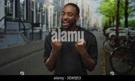 Junger afroamerikanischer Mann mit geballten Fäusten, der auf einer Straße lächelt, die von Fahrrädern und Reihenhäusern gesäumt ist; persönlicher Triumph. Stockfoto