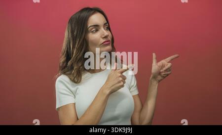Frau zeigt beide Zeigefinger nach außen nach rechts, während sie in einem roten Studio seitwärts schaut; Neugierrichtung fokussiert die Erwartung. Stockfoto