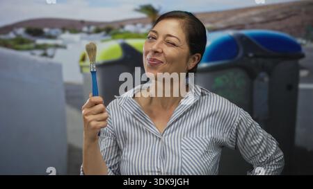 hispanische Frau mittleren Alters hält Pinsel in der Hand, indem sie Abfalleimer auf der Straße recycelt; Kreativität Verspieltheit Zufriedenheit Kunstfertigkeit. Stockfoto